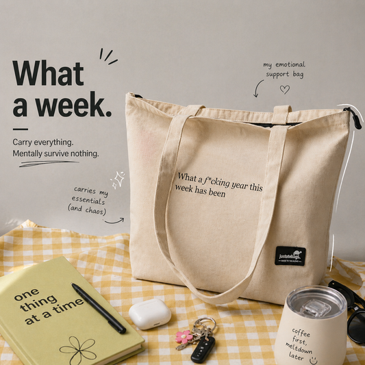 Beige tote bag with humorous text on a checkered tablecloth with notebook, pen, and other items.