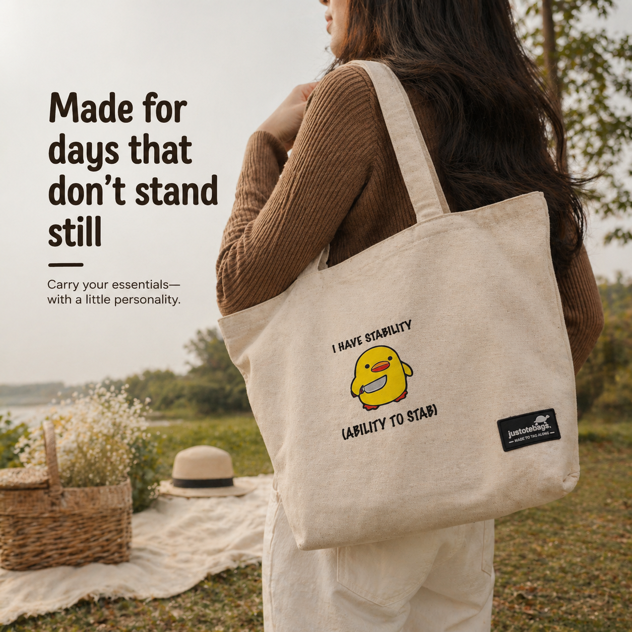 Person holding a beige tote bag with a cartoon character and text, standing outdoors near a picnic setup.
