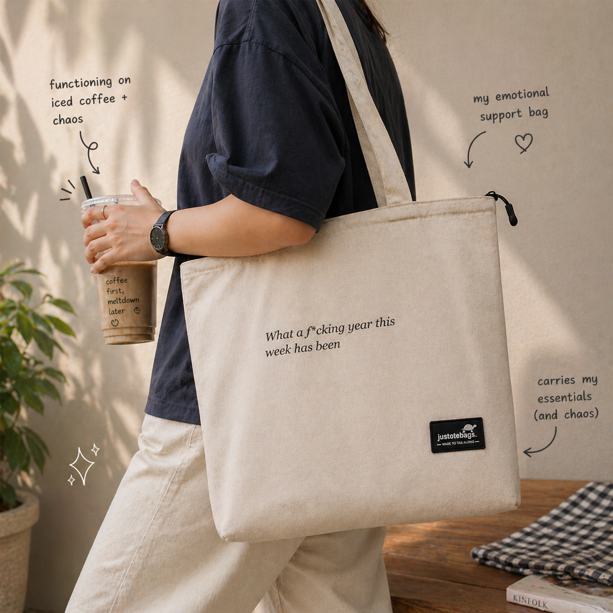 Person holding a beige tote bag with text and a drink, with a neutral background