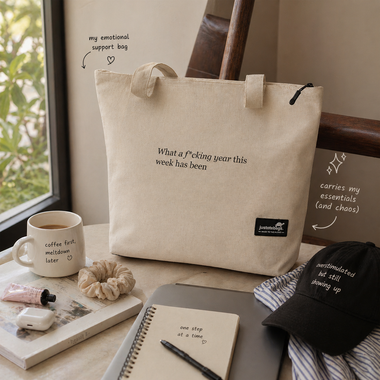 Beige tote bag with text, mug, notebook, and cap on a table near a window.